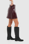 Oxblood Meeghan Mini Skirt in glossy vegan leather, paired with classic black knee-high boots, embodies effortless high-end style for a fun, versatile look.