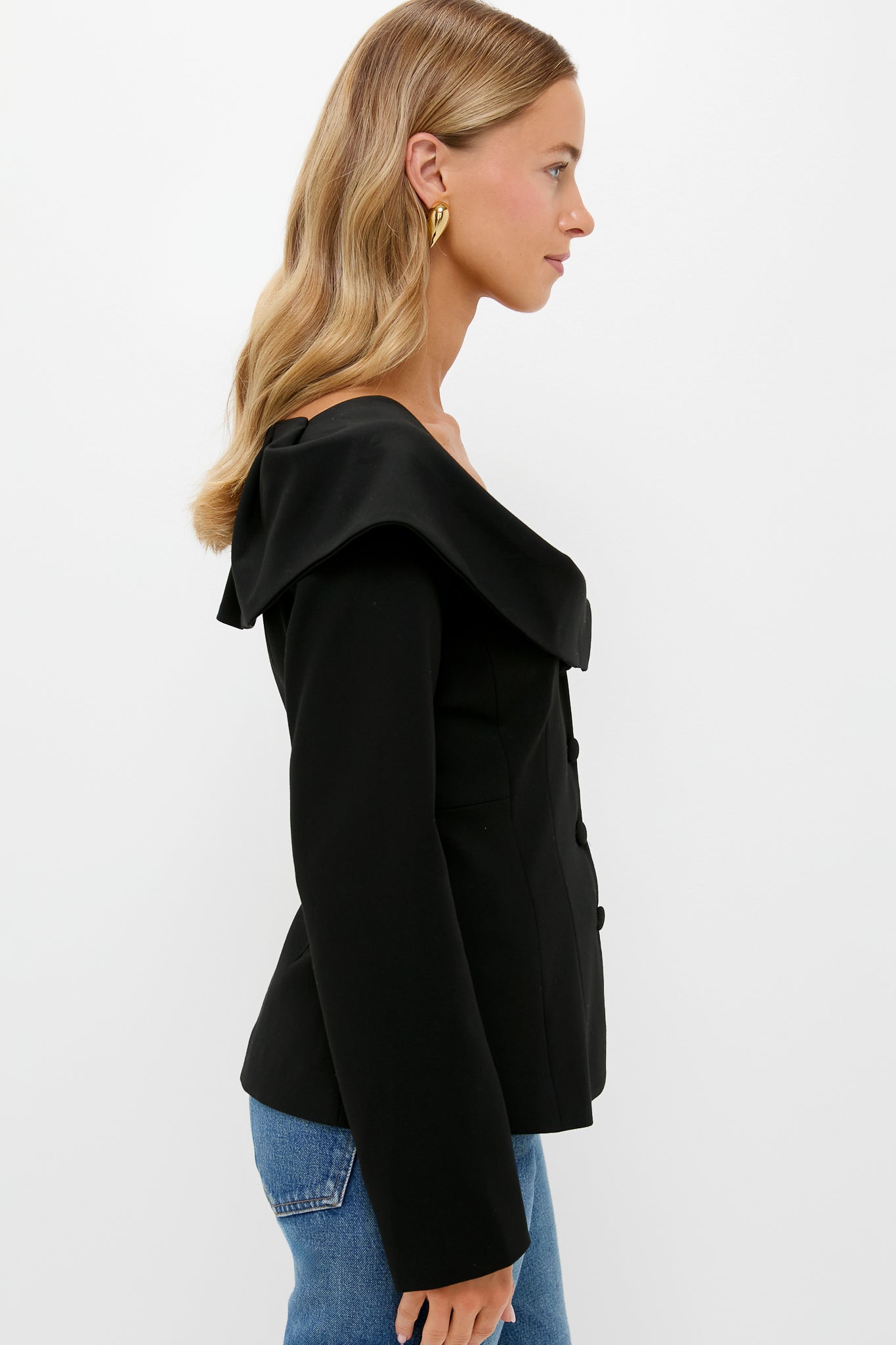 A woman in profile wears a black Tuckernuck Monroe Top with an off-the-shoulder, wide collar design, blue jeans, and gold hoop earrings against a plain white background.