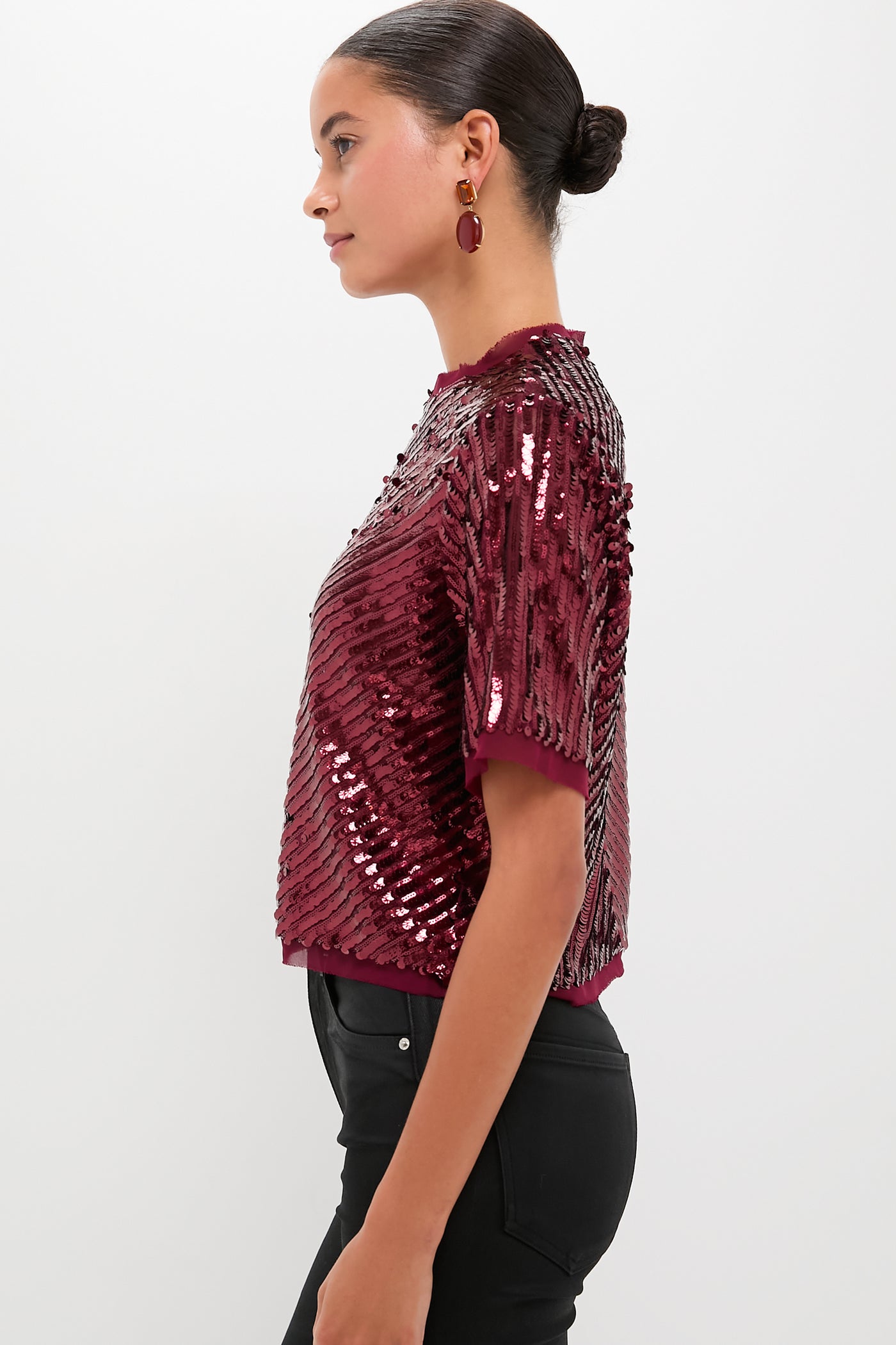 A woman in profile wears a Hunter Bell red sequin top with chiffon trim, black pants, coordinating burgundy earrings, and her hair styled in a neat bun, set against a plain background.