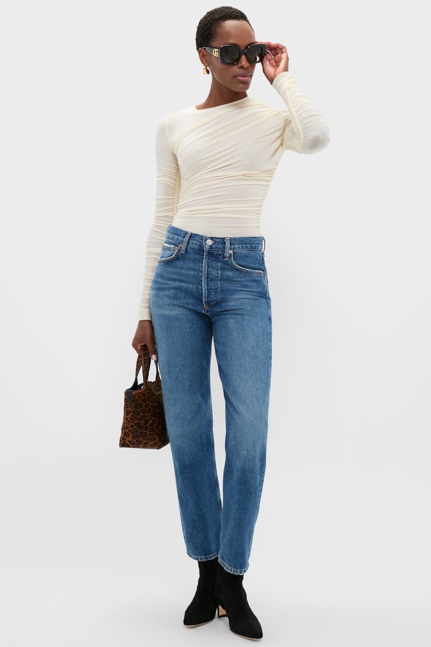 Person wearing a fitted cream ruching top, AGOLDE high-waisted blue jeans, black ankle boots, sunglasses, and carrying a small leopard print handbag.
