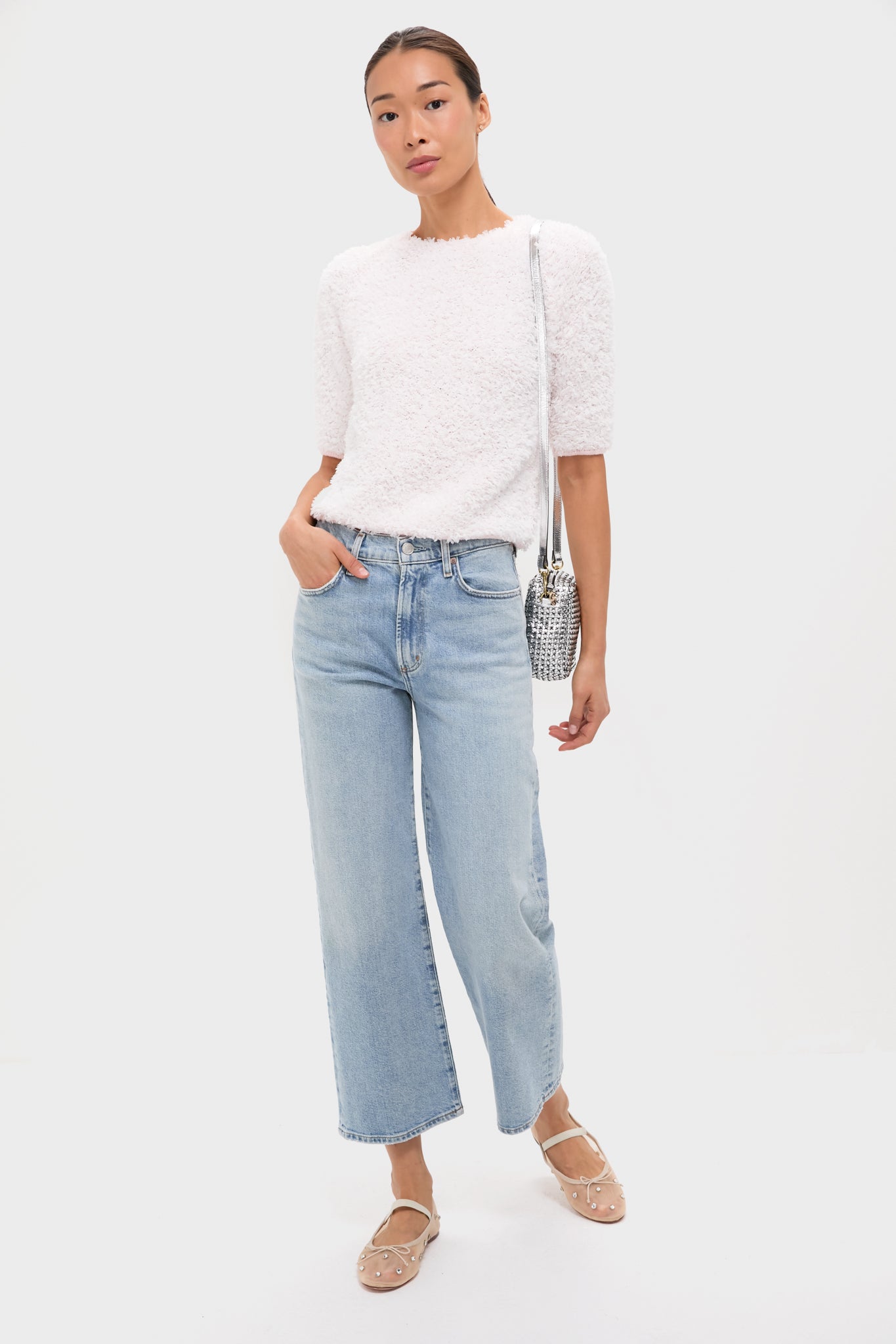 A woman stands against a plain background wearing a white textured short-sleeve top, light blue wide-leg jeans, embellished beige flats, and carries a Clare V handwoven leather crossbody bag with a detachable strap.