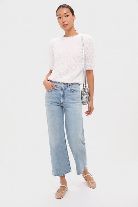 A woman stands against a plain background wearing a white textured short-sleeve top, light blue wide-leg jeans, embellished beige flats, and carries a Clare V handwoven leather crossbody bag with a detachable strap.