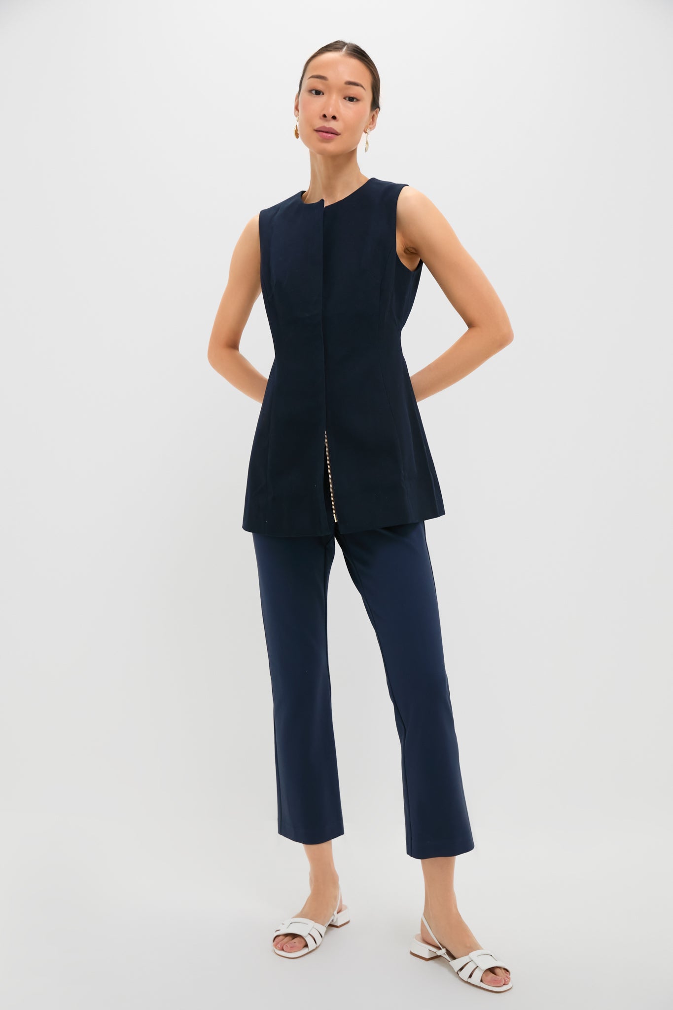 A woman wears a sleeveless navy tunic and Tuckernuck Navy Ponte Ashford Pants with a cropped kick flare hem, paired with white sandals.
