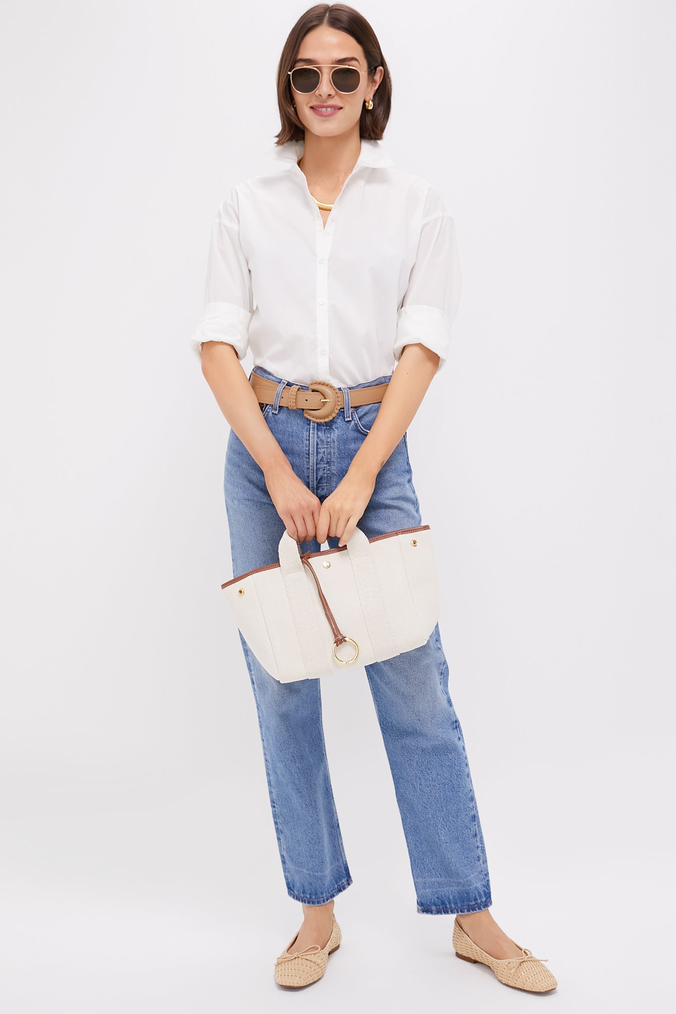 A woman in a Tuckernuck cotton button-down made from wrinkle-resistant fabric, blue jeans, a beige belt and flats, accessorized with sunglasses and a white handbag.