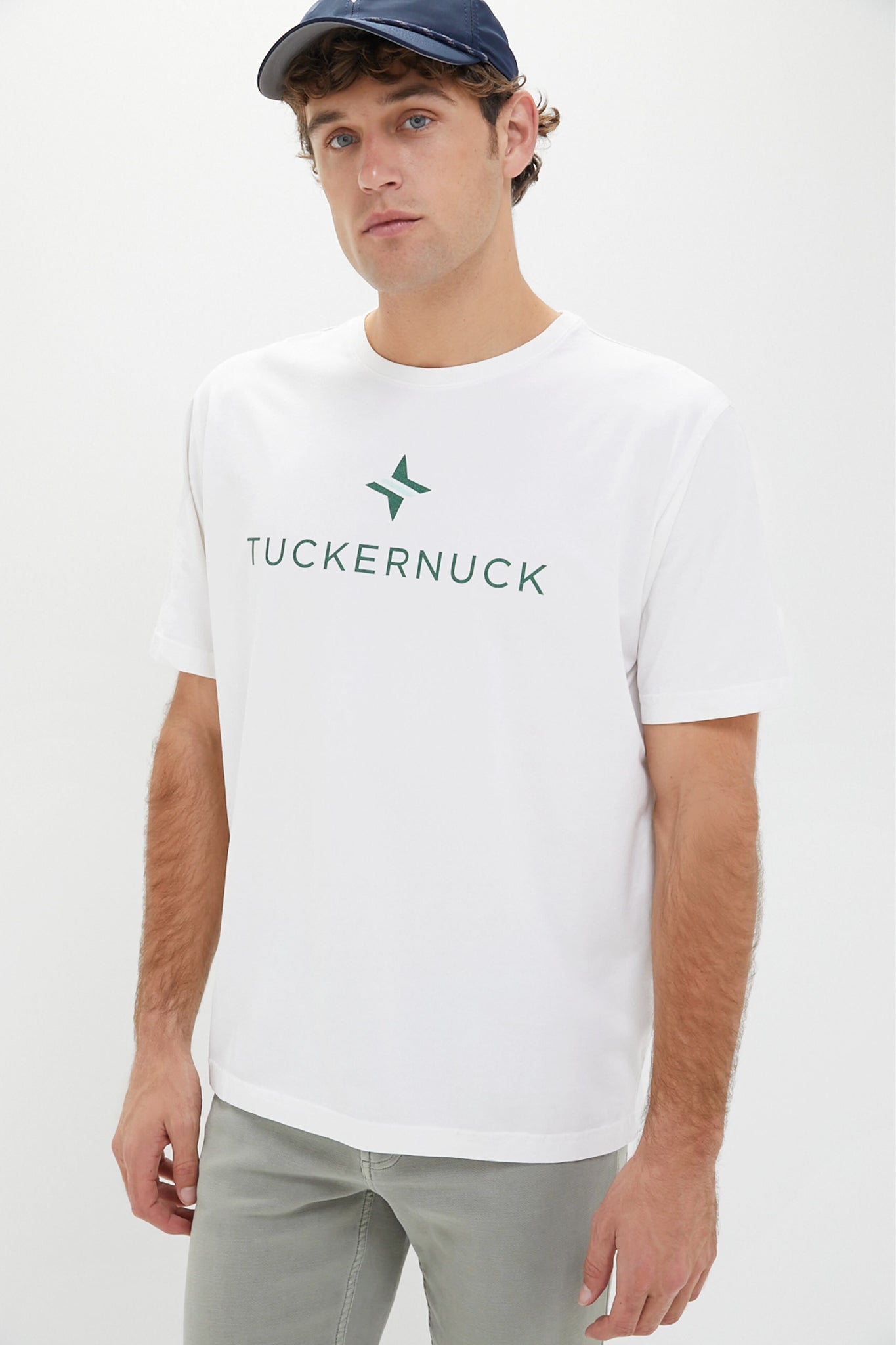 A man wears a Peter Millar White Tuckernuck Tee crafted from performance fabric with UPF 50+ protection, featuring "TUCKERNUCK" and a green logo on the front, paired with a navy cap.