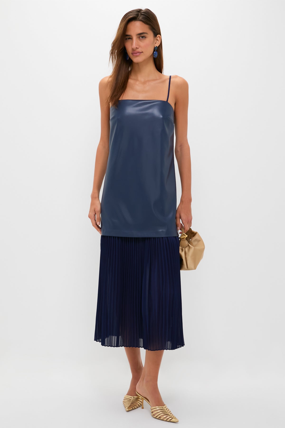 Model wears the Mixed Media Leather Pleated Ozzy Midi Dress, featuring a classic navy sleeveless top and pleated skirt, paired with metallic gold mules and a light tan pouch.