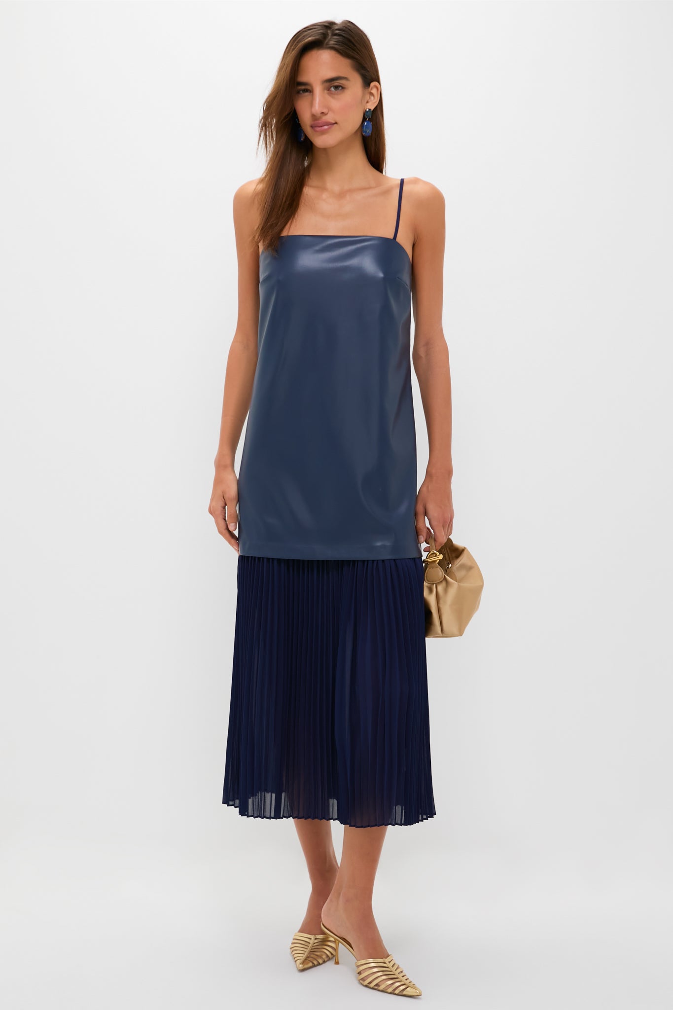 Model wears the Mixed Media Leather Pleated Ozzy Midi Dress, featuring a classic navy sleeveless top and pleated skirt, paired with metallic gold mules and a light tan pouch.