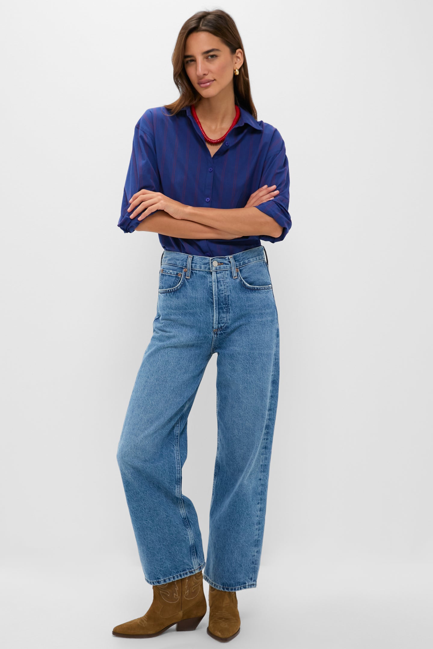 A woman stands with arms crossed in a Tuckernuck women's blouse—a blue 100% cotton button-down shirt—paired with light blue high-waisted jeans and brown ankle boots, set against a plain white background.