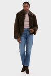 A person stands facing forward in a Stand Studio brown luxury faux fur jacket, beige turtleneck sweater, blue jeans, and brown ankle boots against a plain white background.