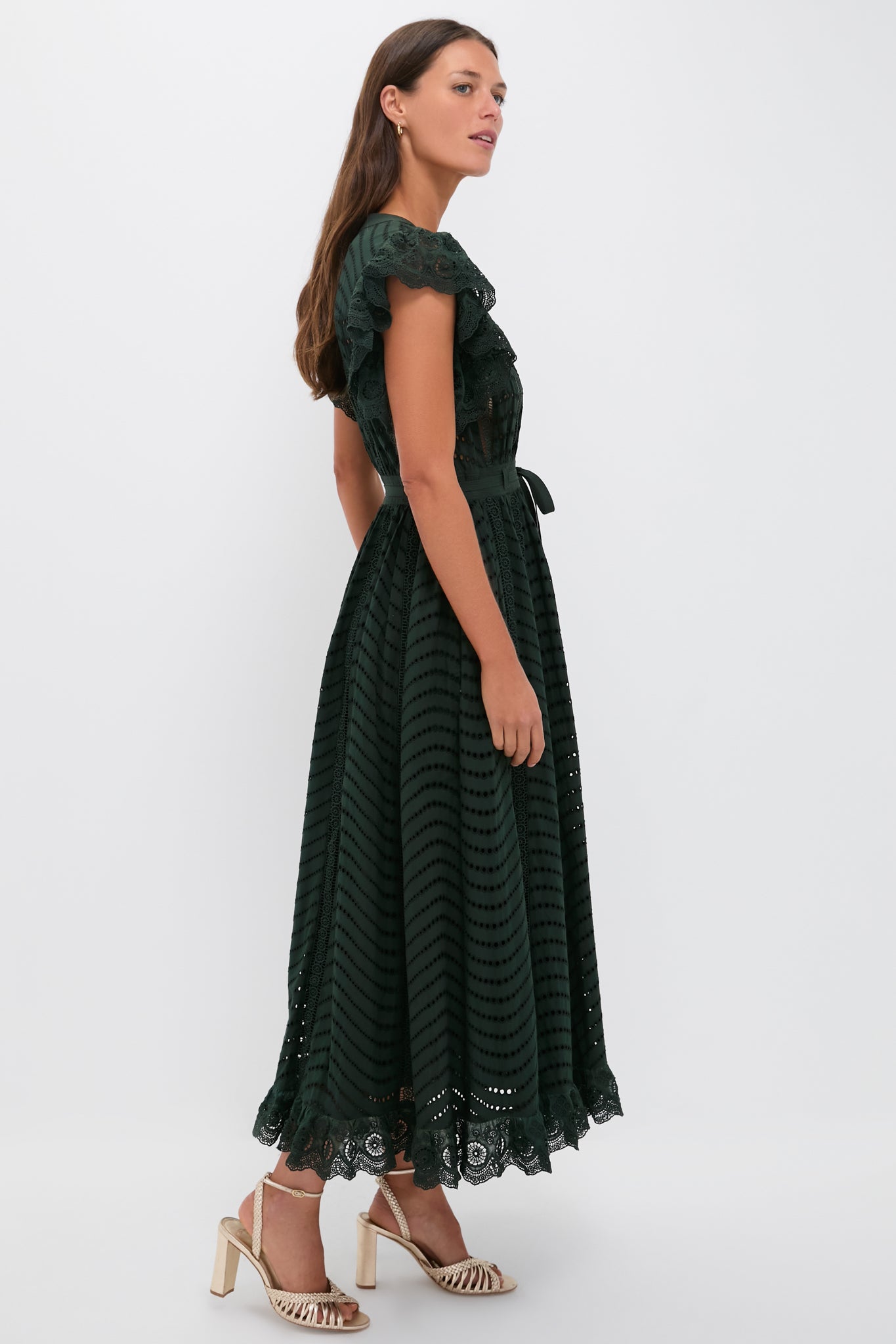 A woman in the Ulla Johnson Olga Dress—a dark green lace cocktail dress with scalloped edges and a belted waist—pairs it with cream strappy heels, standing against a plain white background.