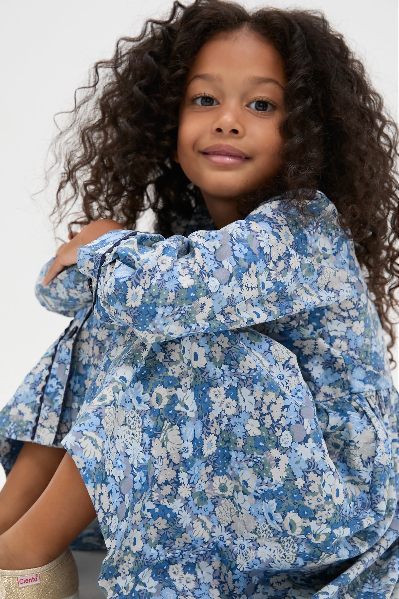 Young child in a classic, long-sleeve floral button-front dress by Rachel Riley, featuring effortless elegance and fun blue floral patterns, paired with high-end metallic-gold Cienta shoes.
