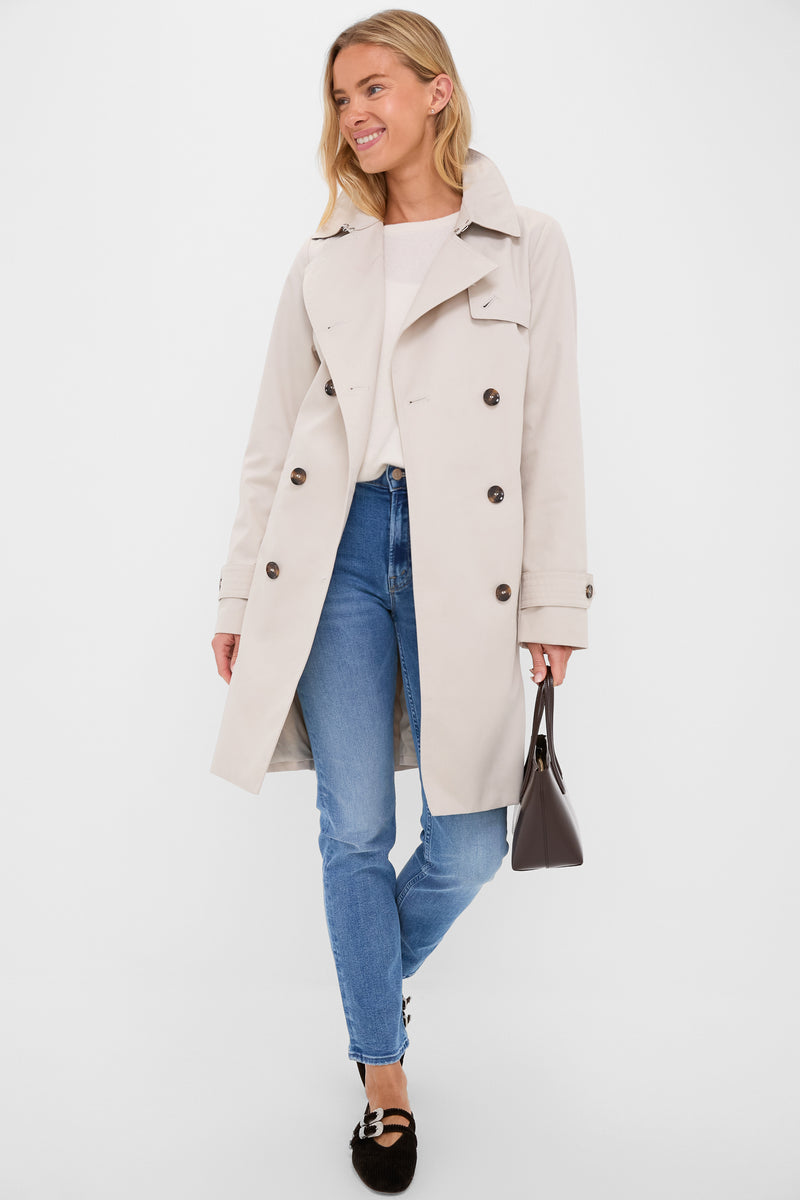 Young woman in a classic Mist Short Greta Showerproof Jacket, showcasing an effortless, high-end style with double-breasted design and fun details, paired with jeans and a handbag.