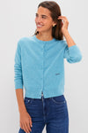 A woman smiles with one hand touching her hair, wearing a Tuckernuck light blue cardigan and dark blue jeans.
