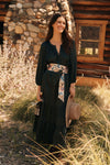 Woman standing in front of a cabin in a long green dress with a floral belt. 