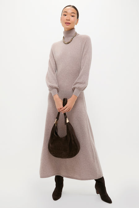 A person in a Tuckernuck light gray cashmere sweater dress with an A-line silhouette, dark boots, a chunky necklace, and holding a dark brown suede handbag stands against a plain white background.