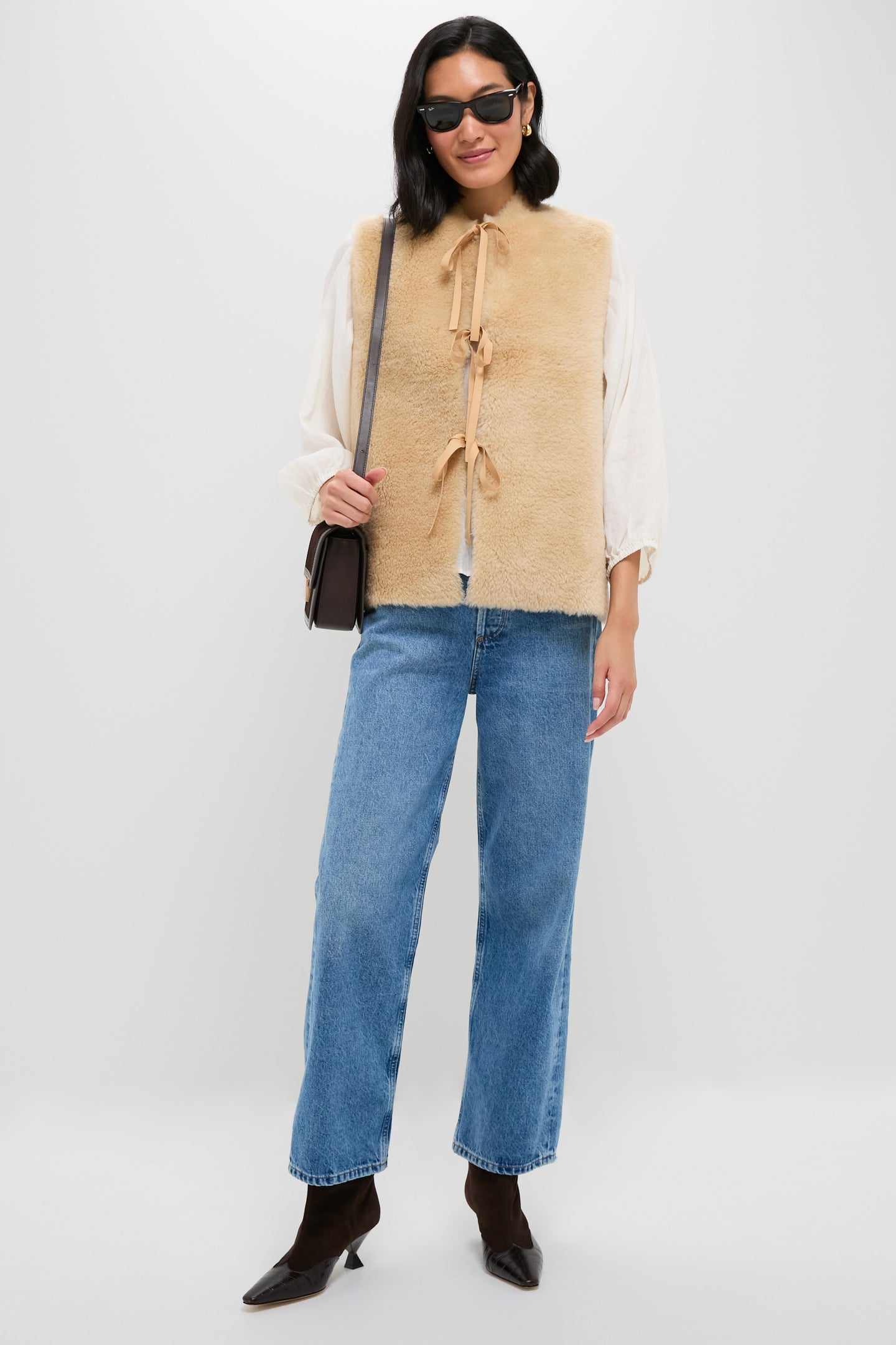 A person in sunglasses, blue jeans, brown boots, and a cream blouse layered with the Sea New York beige wool reversible vest with ribbon ties, carrying a brown shoulder bag, stands against a white background.