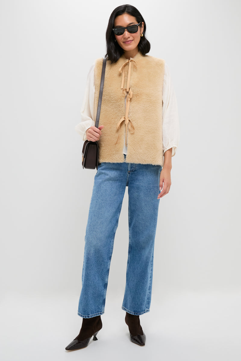 A person in sunglasses, blue jeans, brown boots, and a cream blouse layered with the Sea New York beige wool reversible vest with ribbon ties, carrying a brown shoulder bag, stands against a white background.