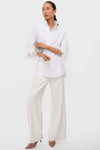 A woman stands against a plain background wearing a Tuckernuck feathered blouse with a stand collar and button front, wide-leg white pants, nude heels, and holding a small clutch.