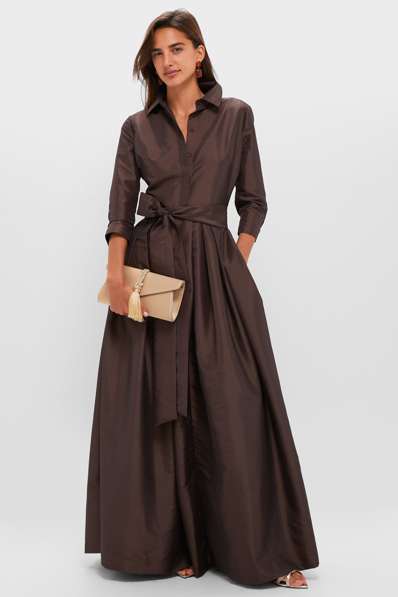 Model showcasing the classic Champagne Satin Gabrielle Clutch with braided tassel, adding an effortless high-end flair to her fun, voluminous brown shirt-style dress ensemble.