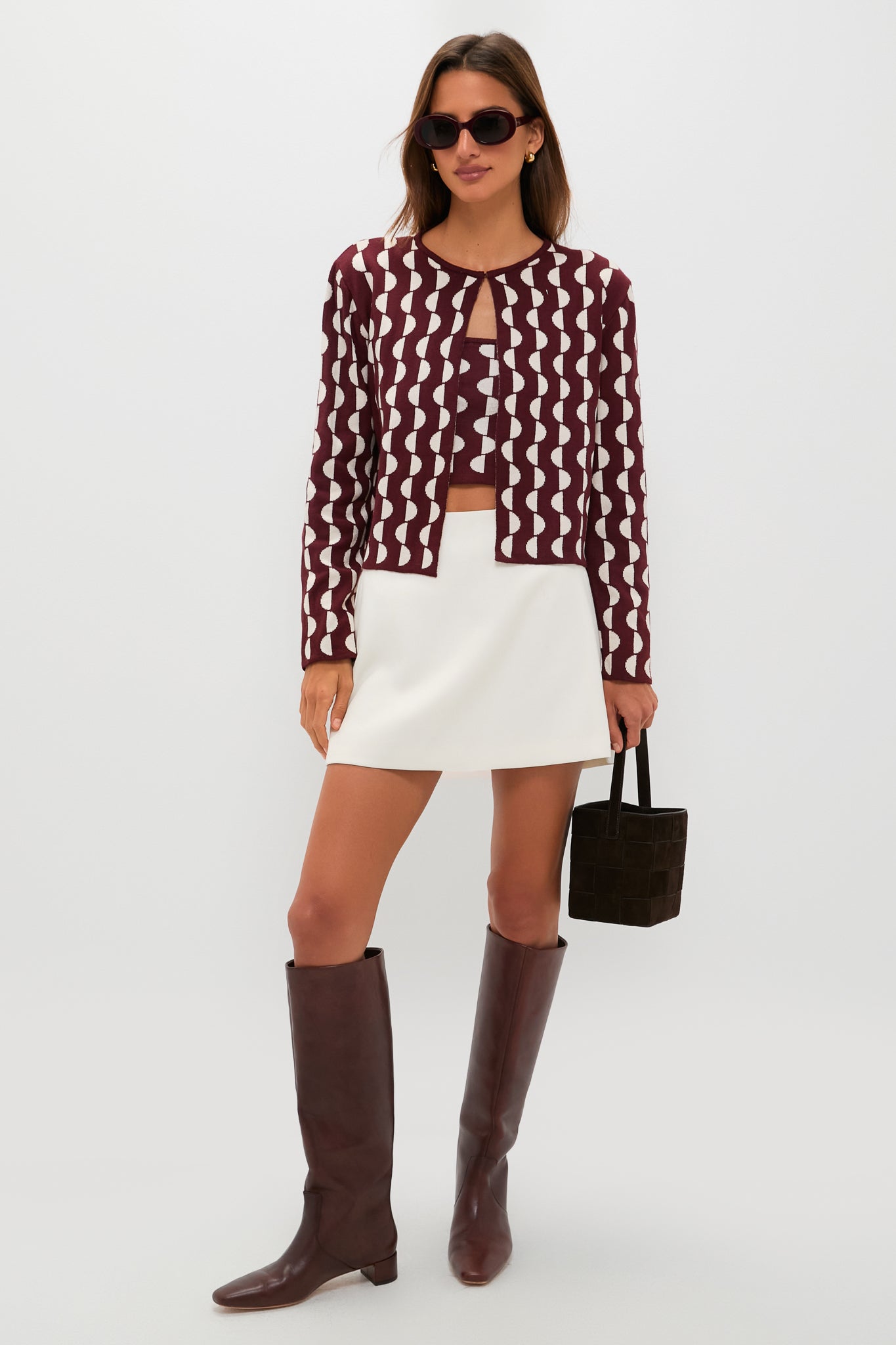 A woman styles a Dress To maroon and white patterned cardigan over a matching top, paired with a white mini skirt, brown knee-high boots, sunglasses, and a brown handbag.