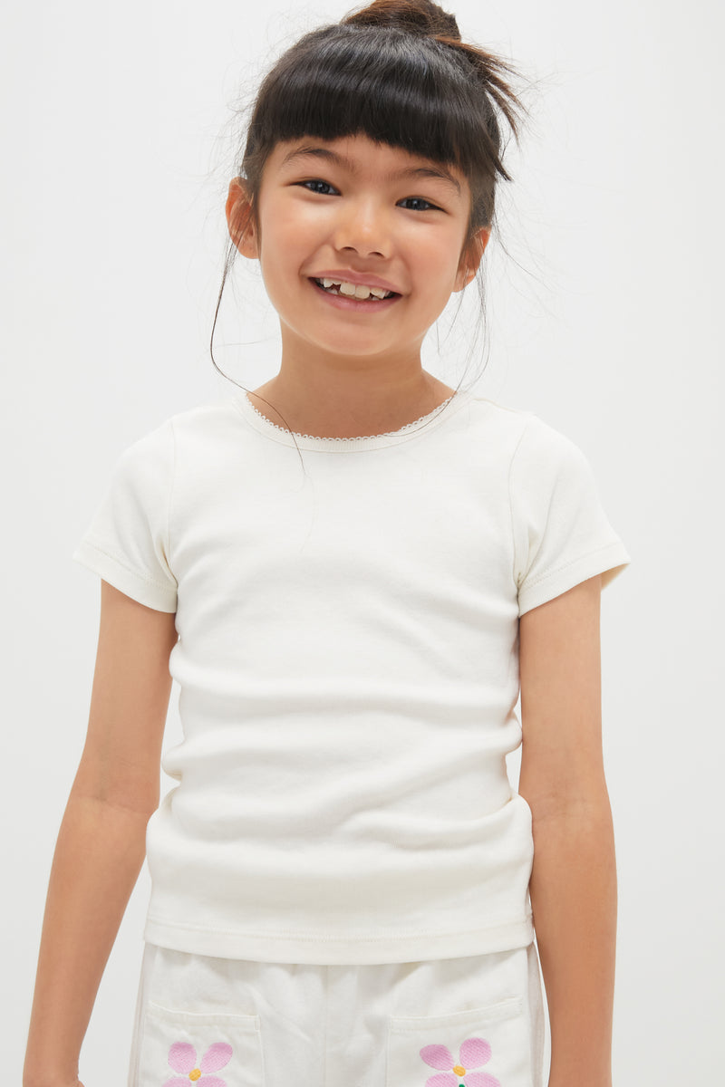 A young girl with dark hair in a bun wears a Pink Chicken short sleeve 100% cotton crewneck t-shirt and white shorts with pink flower designs, standing against a plain light background.