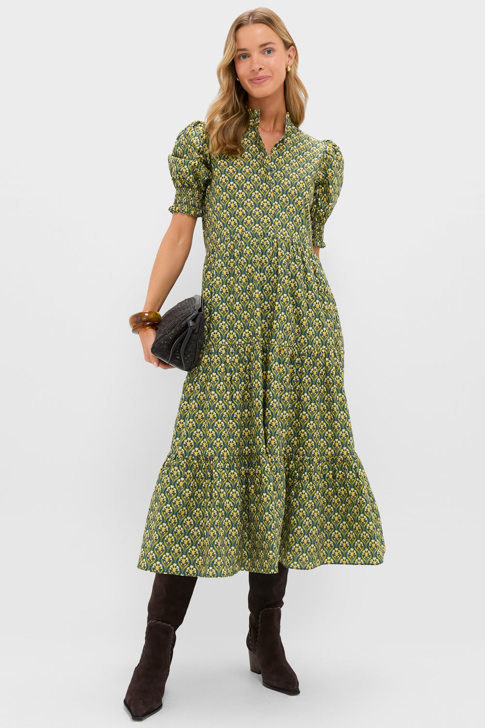 A woman wears a Hyacinth House green floral print midi dress with short puff sleeves and a tiered skirt, paired with black knee-high boots and a black clutch bag.