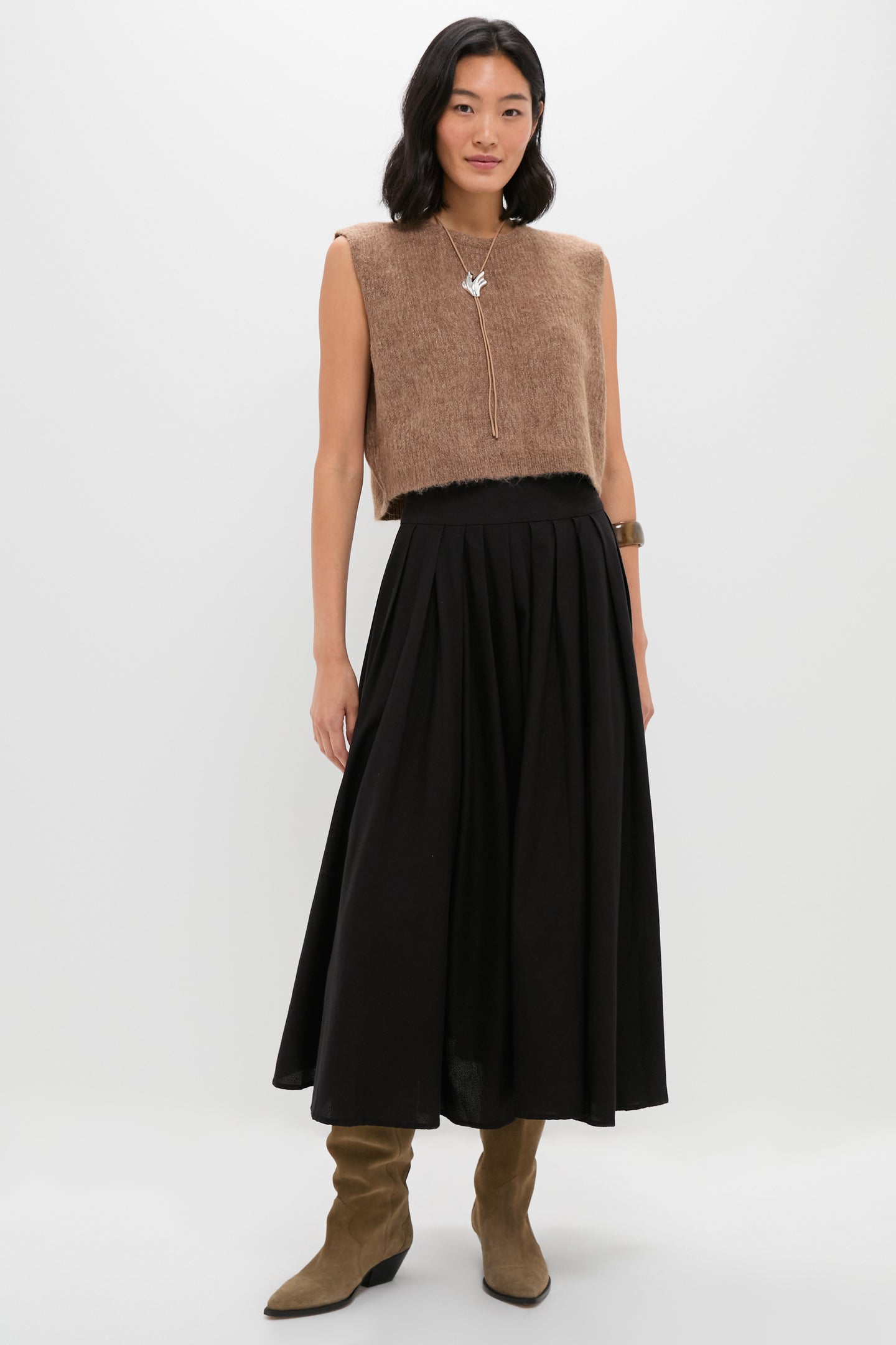 A woman wears THE GREAT. sleeveless brown fuzzy top, long black pleated skirt with a wide waistband, brown suede boots, necklace, and bracelet against a plain white background.