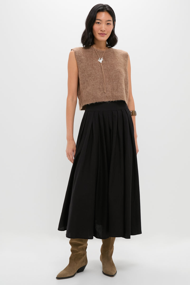 A woman wears THE GREAT. sleeveless brown fuzzy top, long black pleated skirt with a wide waistband, brown suede boots, necklace, and bracelet against a plain white background.