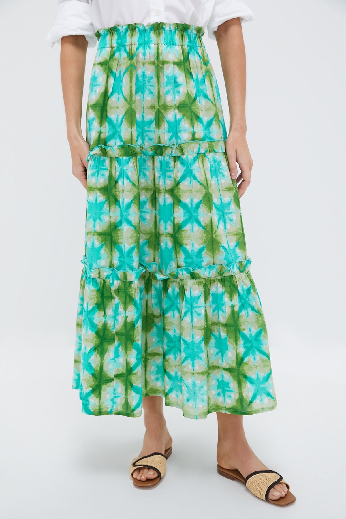 A person wearing a Tnuck Beach white shirt and long tiered skirt with green and blue tie-dye layers, an elastic ruffle waistband, paired with tan slide sandals.