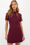 A woman wears a Hyacinth House burgundy lace mini dress with short sleeves and a delicate overlay, standing in a simple setting.