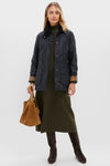A woman wearing a Barbour Beadnell Wax Jacket over an olive green sweater dress, carrying a brown suede handbag, and wearing knee-high dark brown boots by Barbour.