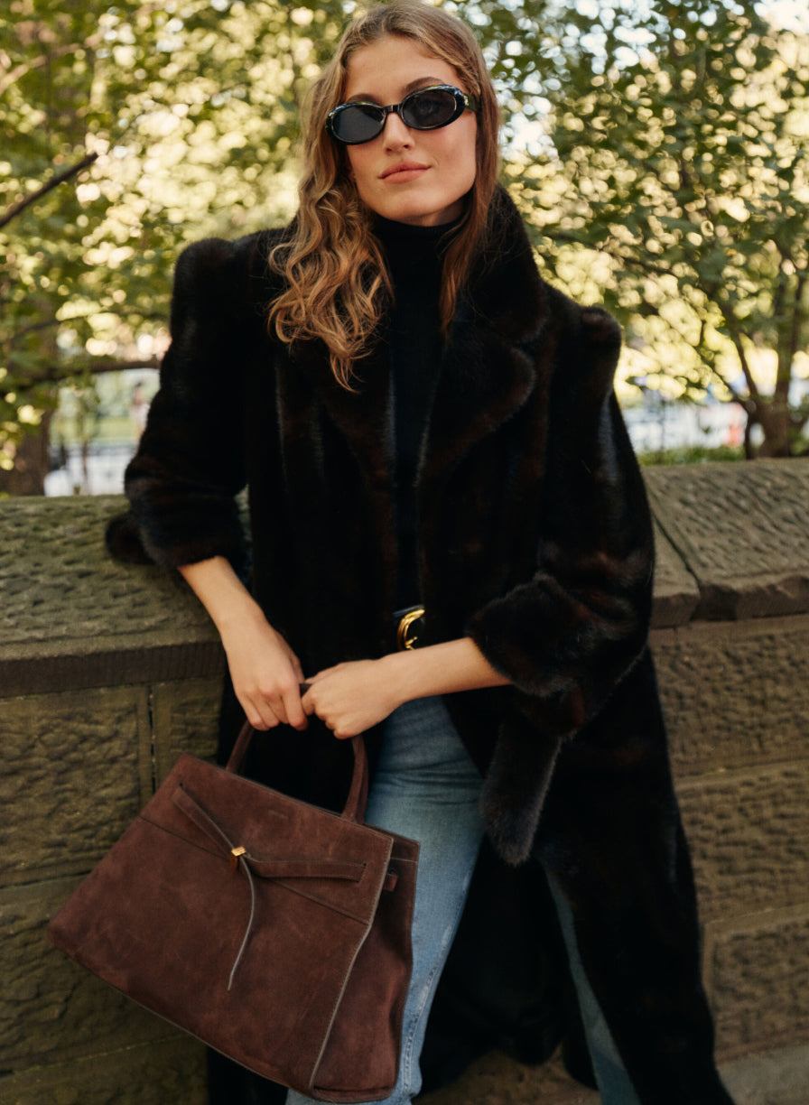 A woman leans on a city park bridge wearing a full-length fur coat, a black turtleneck, and a pair of blue jeans while carrying a brown suede tote bag and wearing sunglasses.