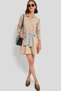 A woman wearing a Tuckernuck beige classic pique polo dress with a striped sweater at her waist, brown loafers, sunglasses, and a black shoulder bag stands against a light gray background.