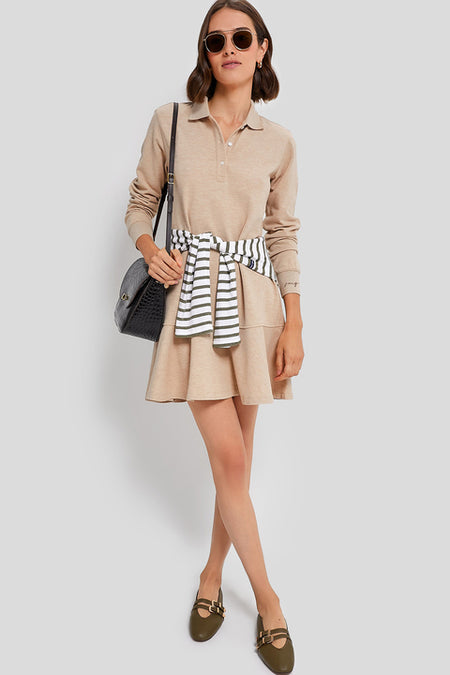A woman wearing a Tuckernuck beige classic pique polo dress with a striped sweater at her waist, brown loafers, sunglasses, and a black shoulder bag stands against a light gray background.