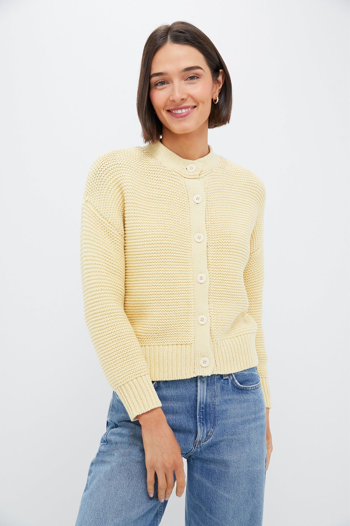 A woman with short brown hair stands in a well-lit space wearing blue jeans and a light yellow chunky knit cardigan by Alex Mill.