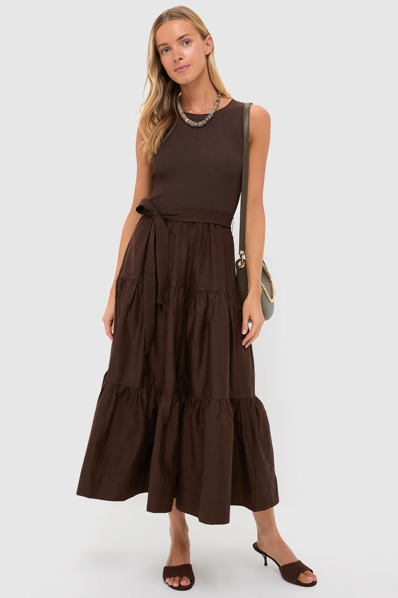 A woman stands against a plain background wearing a sleeveless brown belted maxi dress with a tiered skirt, Schutz heeled sandals, a necklace, and a shoulder bag.