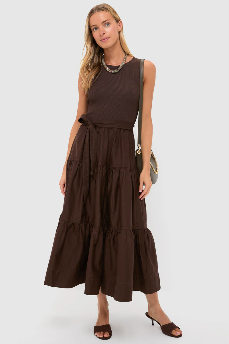 A woman stands against a plain background wearing a sleeveless brown belted maxi dress with a tiered skirt, Schutz heeled sandals, a necklace, and a shoulder bag.