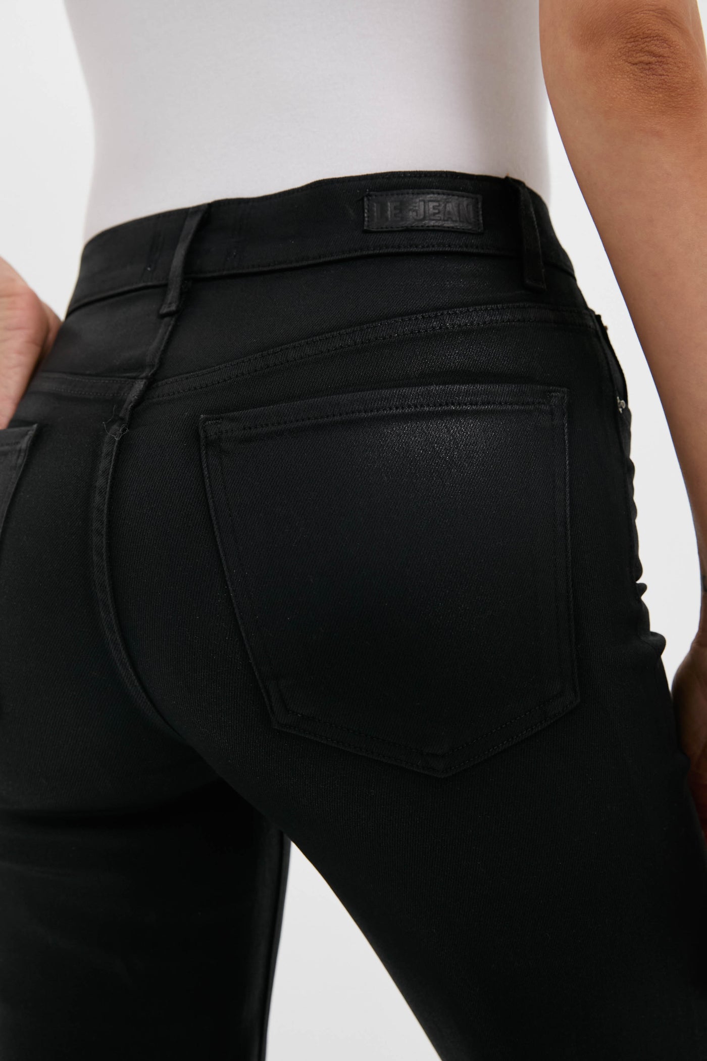 Close-up of a person wearing LE JEAN high rise black coated denim jeans and a white shirt, highlighting the back pocket and waistband label—classic Los Angeles denim style.