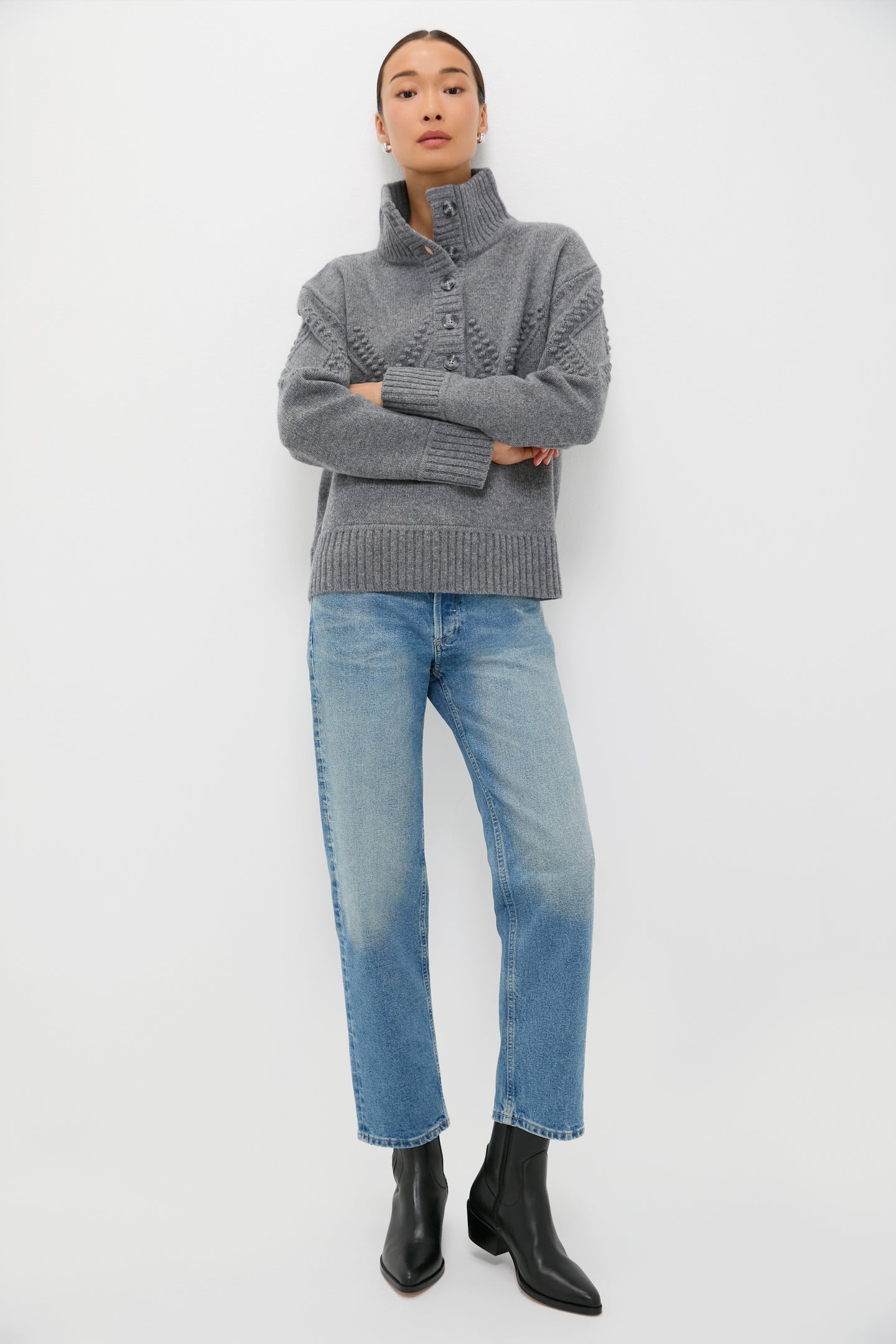 A person stands against a white background wearing the Tuckernuck Gray Henry Sweater in 100% wool, paired with light blue jeans and black ankle boots, arms crossed.