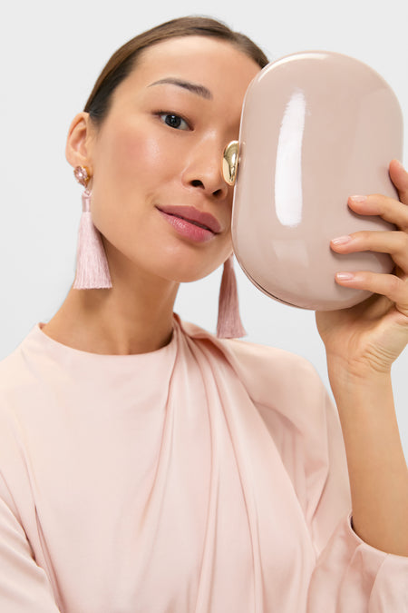 Young woman holds the classic Latte Patent Selena Clutch Bag, an effortless, high-end, glossy nude-beige oval clutch, complementing her fun, minimal style with tassel earrings.