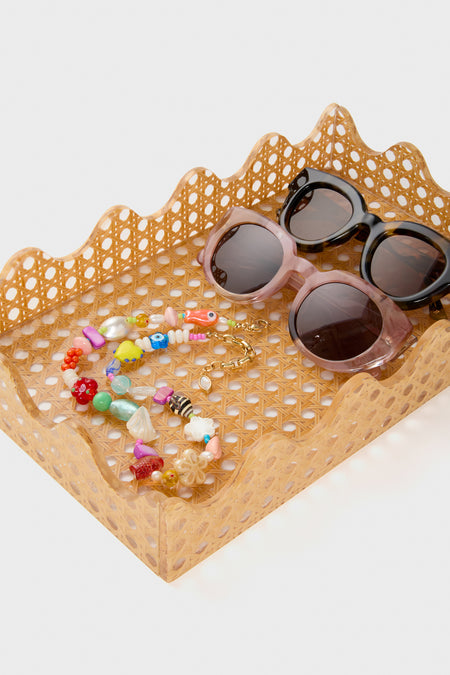 A Tuckernuck Home rectangular acrylic tray with scalloped edges and a rattan interior displays two pairs of sunglasses (black and pink) and a colorful beaded bracelet.