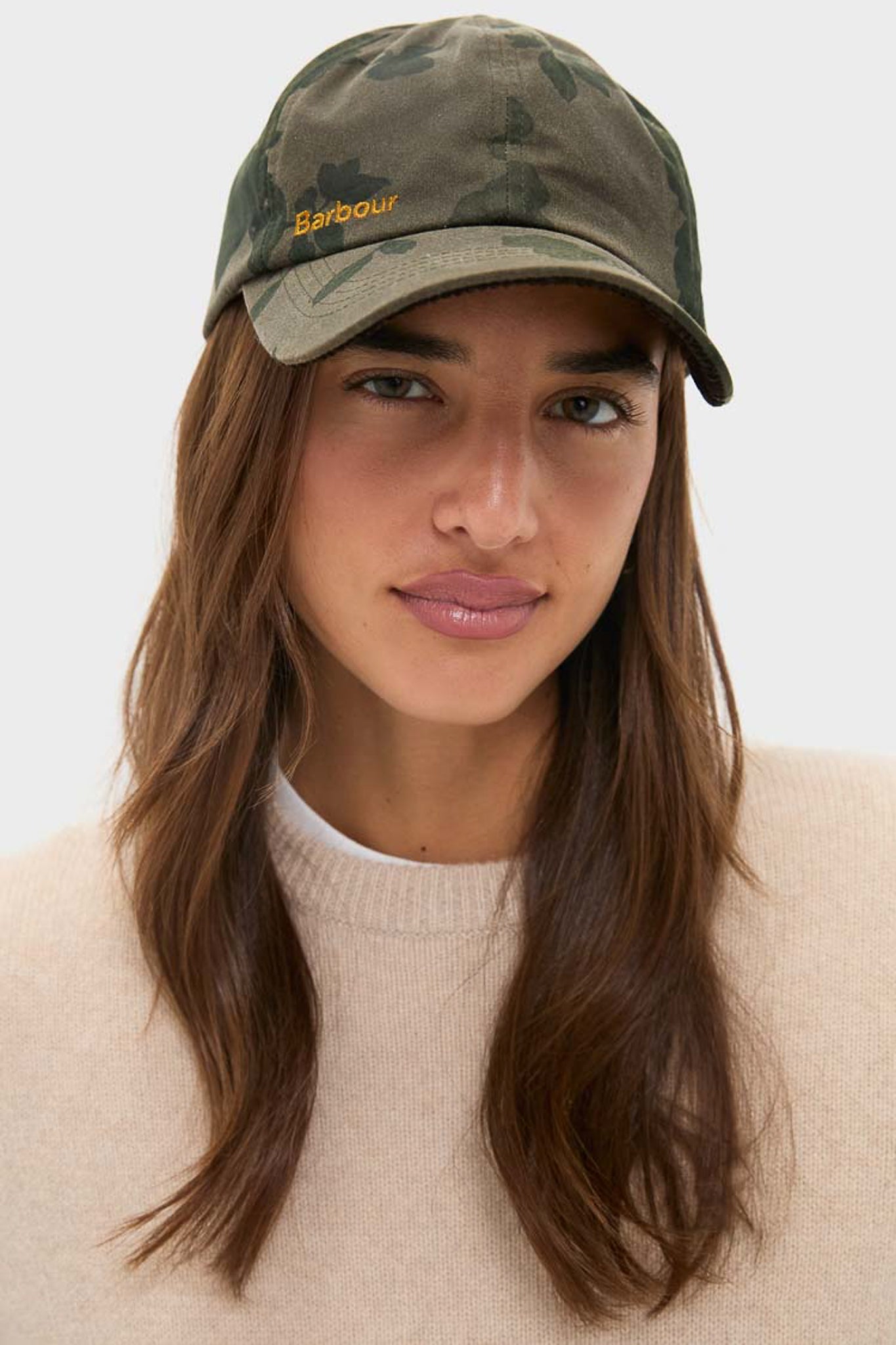 A woman wears a Barbour x Tuckernuck wax cotton camouflage cap and a beige sweater, looking at the camera with a neutral expression.