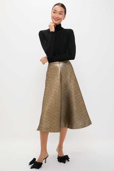 A woman stands against a white background wearing a Tuckernuck black turtleneck sweater, metallic gold circle midi skirt, and black heeled mules with bows.