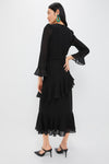 Woman in Black Chiffon Sasha Dress with tiered ruffles, flared sleeves, and a self-tie belt, styled with slingback heels and statement earrings for a classic, high-end look.