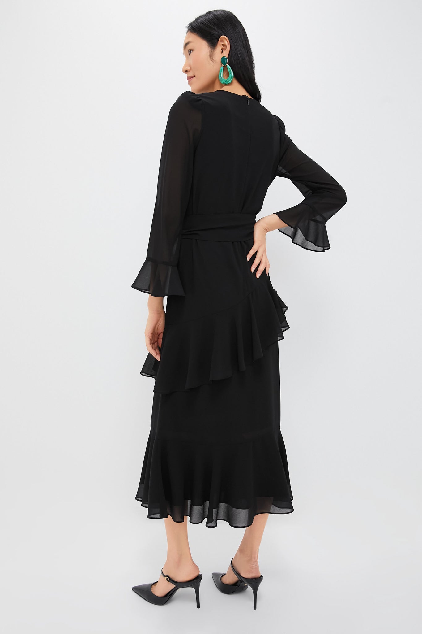 Woman in Black Chiffon Sasha Dress with tiered ruffles, flared sleeves, and a self-tie belt, styled with slingback heels and statement earrings for a classic, high-end look.