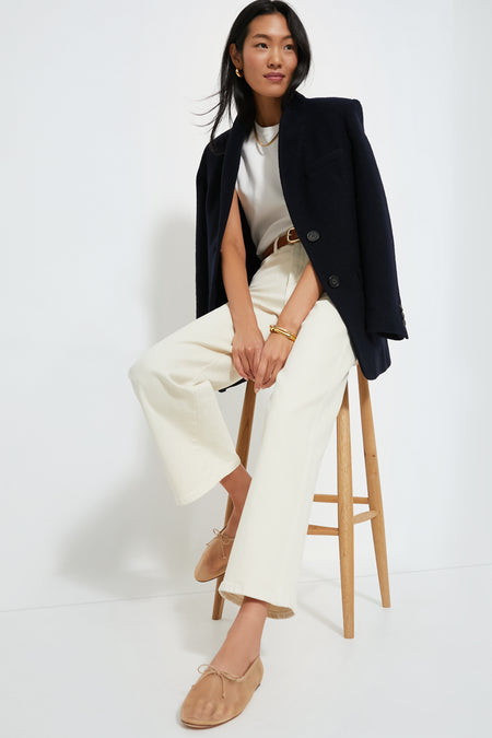A woman sits on a wooden stool, blending masculine and feminine style in a Vanessa Bruno blazer draped over her shoulders, a white shirt, white wide-leg pants, and tan flats.
