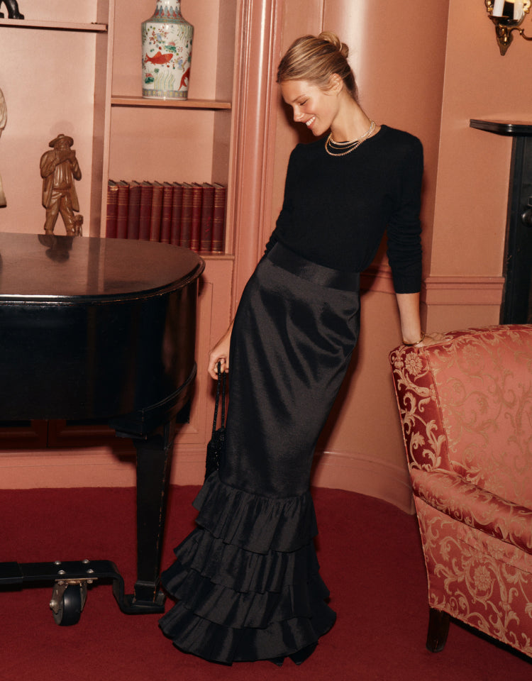 Woman standing by a red chair in a black sweater and long satin skirt with ruffles at the bottom. 