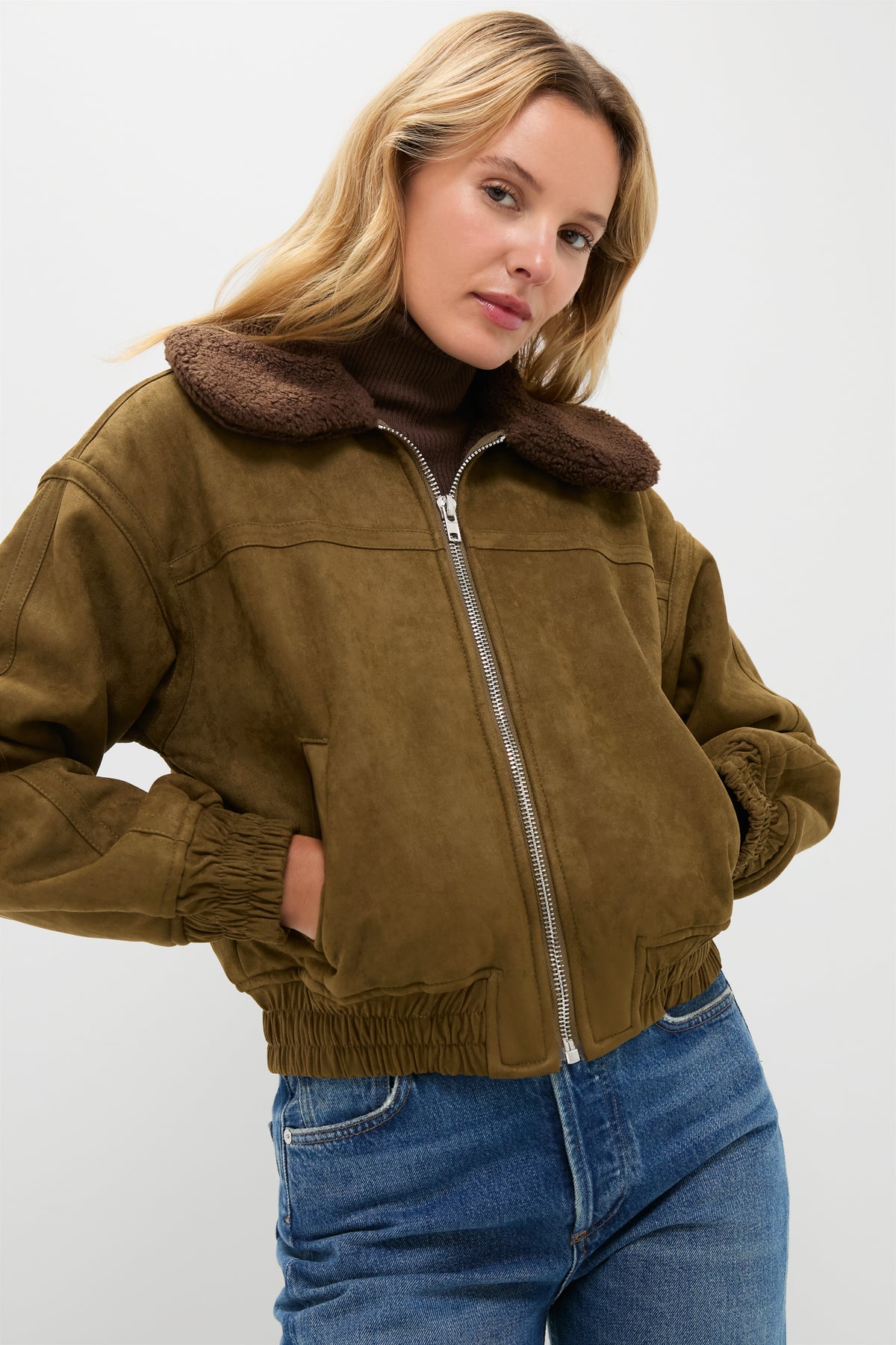 A woman wears a Moon River brown suede bomber jacket with a faux-fur collar, paired with a brown turtleneck and blue jeans.