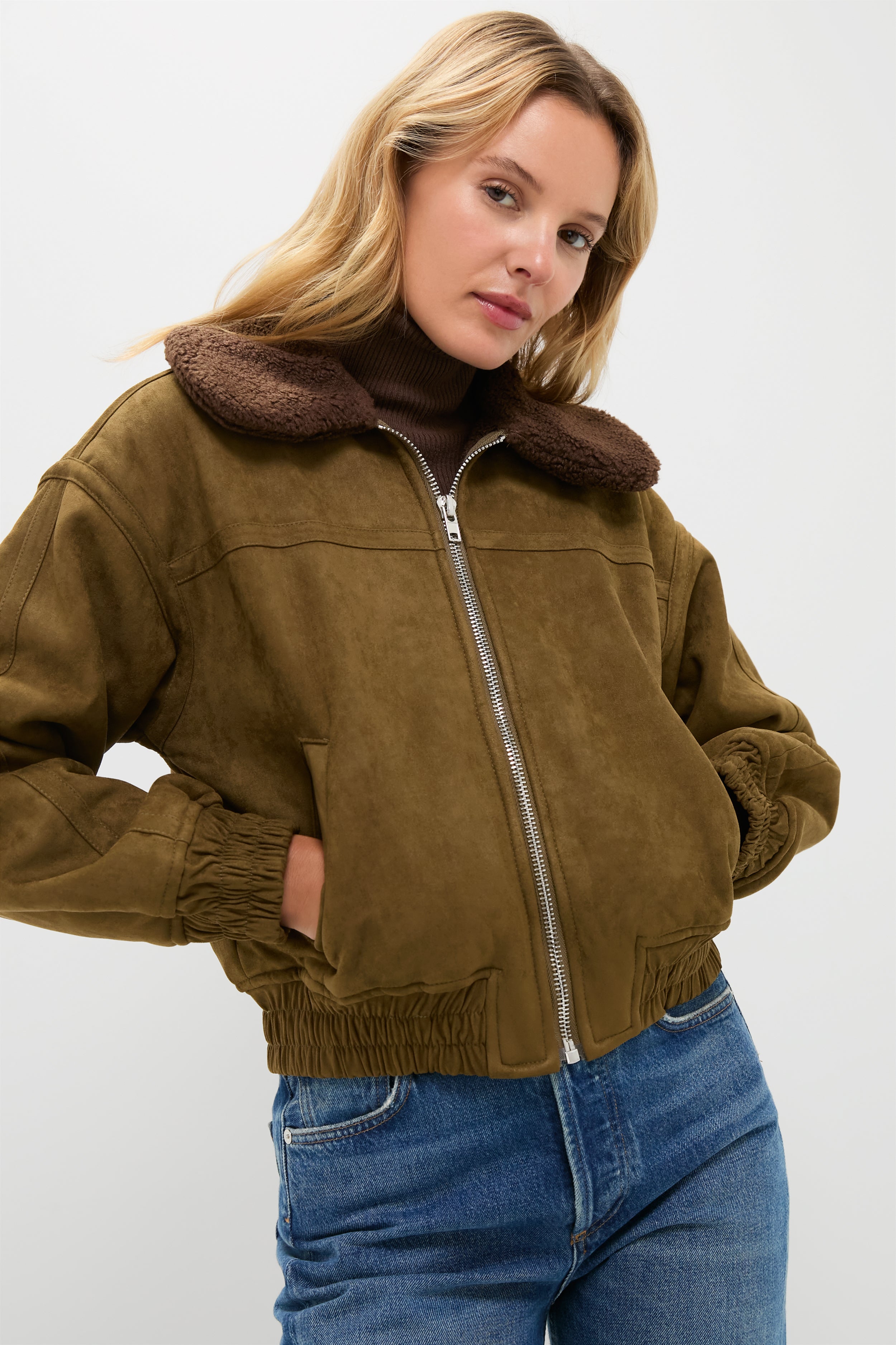 A woman wears a Moon River brown suede bomber jacket with a faux-fur collar, paired with a brown turtleneck and blue jeans.
