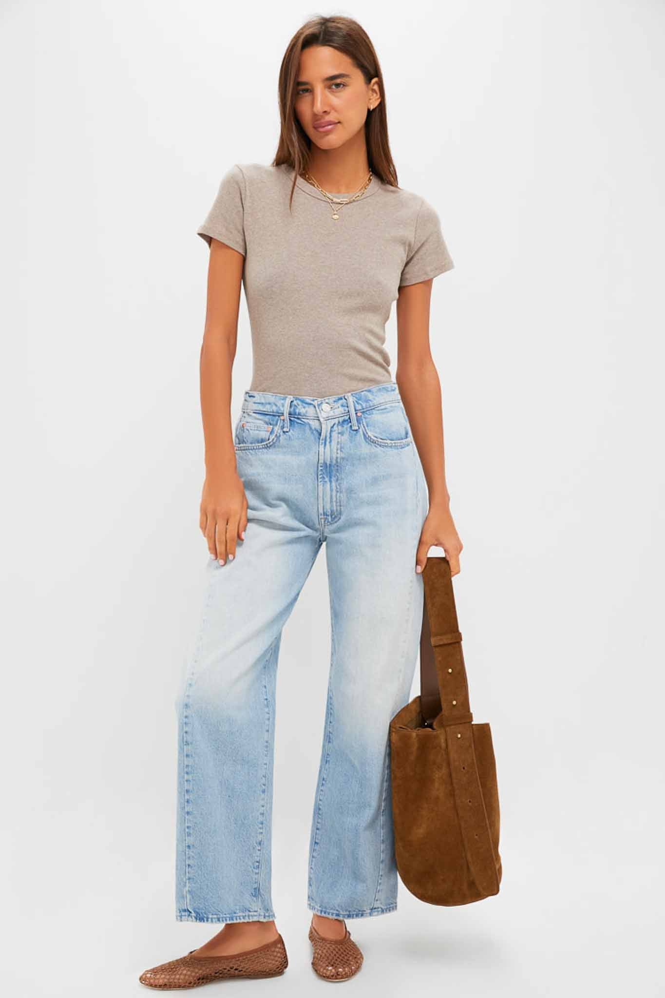 A woman in a beige tee, MOTHER high-rise barrel leg jeans, woven brown flats, and holding a large brown suede bag stands against a plain background.